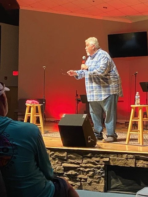 A man in a blue plaid shirt holding a microphone speaking on stage, with two wooden stools, a bottle of water, and a pink cloth on one stool, in a room with a screen and red lighting