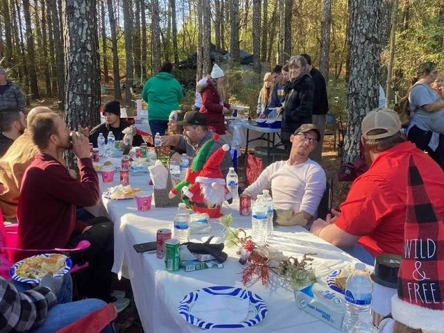Outdoor holiday gathering in a forest with people sitting at a long table decorated with Christmas decorations, holiday plates, and drinks, with some standing around in the background.