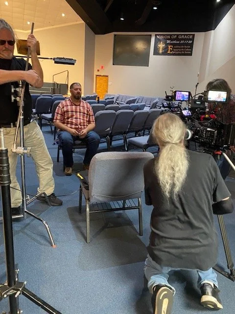 Man in plaid shirt sitting in an empty church or auditorium while crew sets up filming equipment for an interview or recording session.