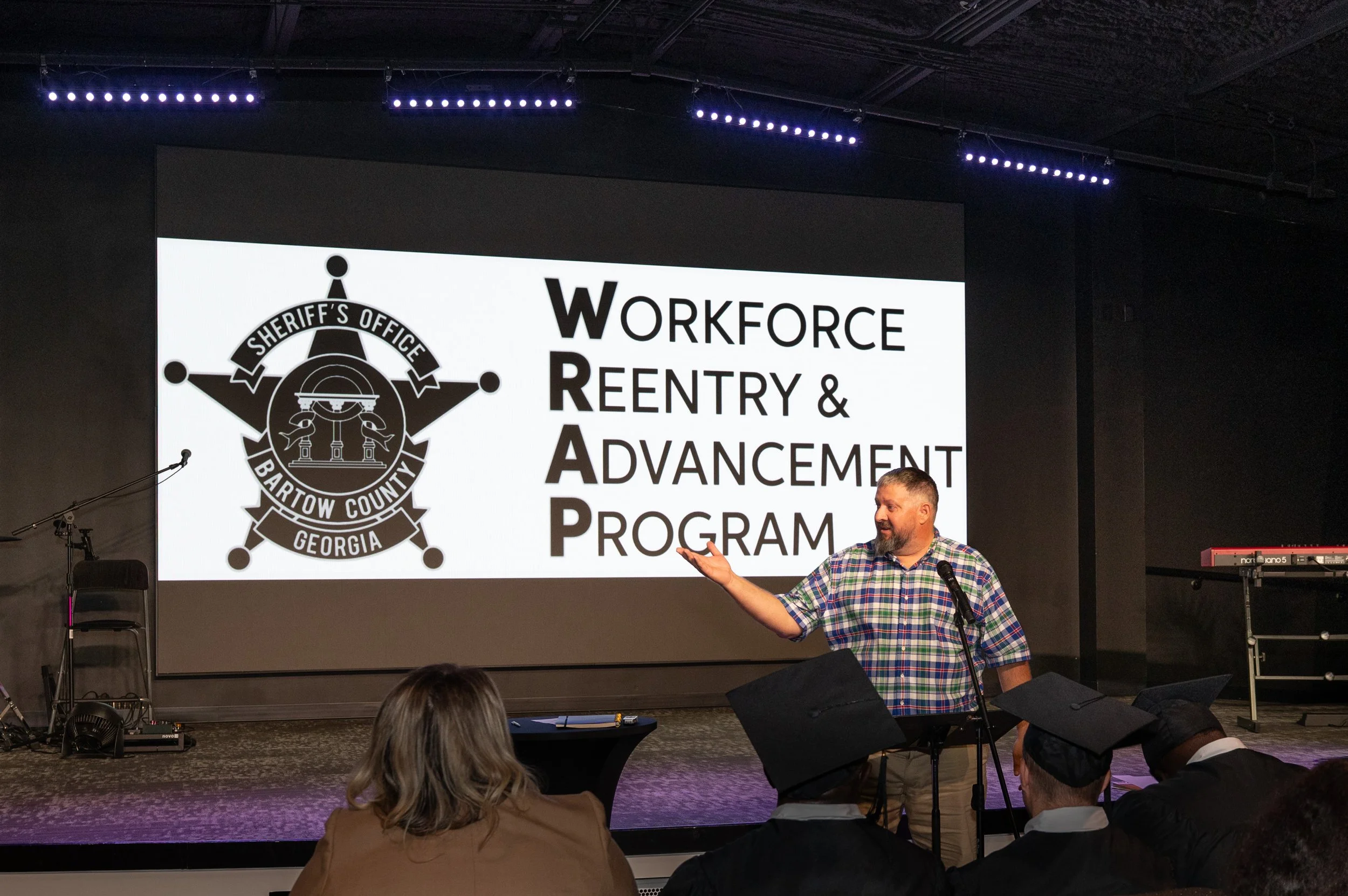 Man giving a presentation at a conference, with a large screen behind him displaying the logo and name of 'Workforce Reentry & Advancement Program' associated with the Sheriff’s Office of Bartow County, Georgia. Several audience members, including so