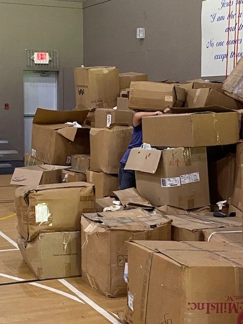 A person hiding behind a stack of large cardboard boxes in a room with wooden floors and gray walls, partially visible with only an arm and part of the body showing.