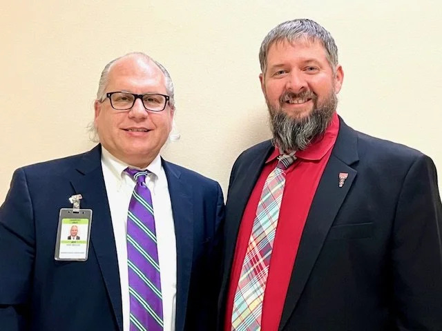 Two men in suits standing against a plain wall, smiling at the camera.