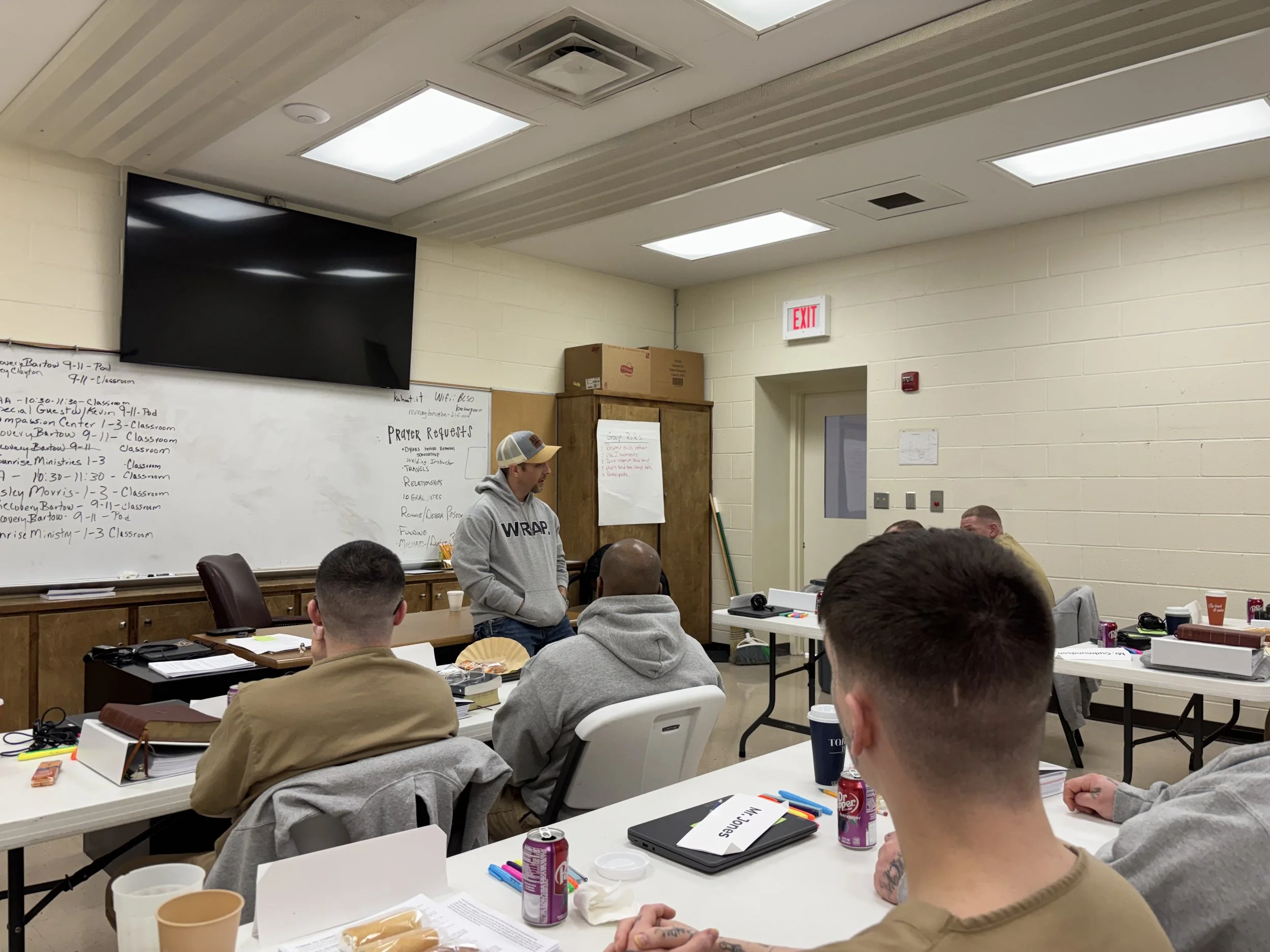 A classroom filled with students seated at tables, listening to a male instructor who is standing near a whiteboard. The whiteboard has handwritten notes, and a large TV is mounted on the wall above it. The room has a drop ceiling with fluorescent lights and an exit sign above a door in the background, with various snacks, drinks, and supplies on the tables.