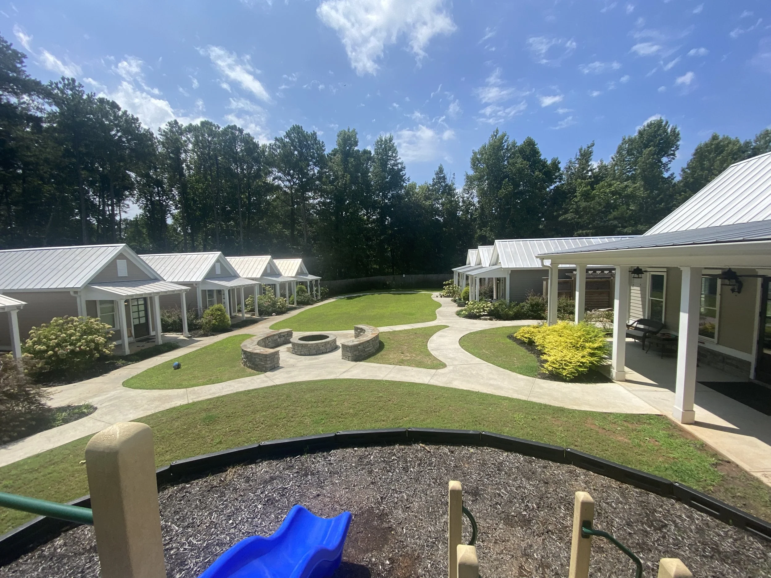 View of a residential neighborhood with small houses, a grassy courtyard with a fire pit, walking paths, and a playground in the foreground. Forest trees in the background under a partly cloudy blue sky.