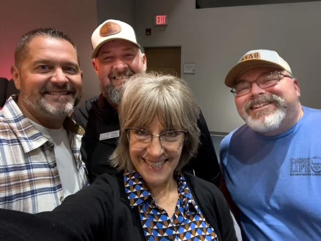 Four smiling adults taking a selfie indoors, with a woman in the front and three men behind her.
