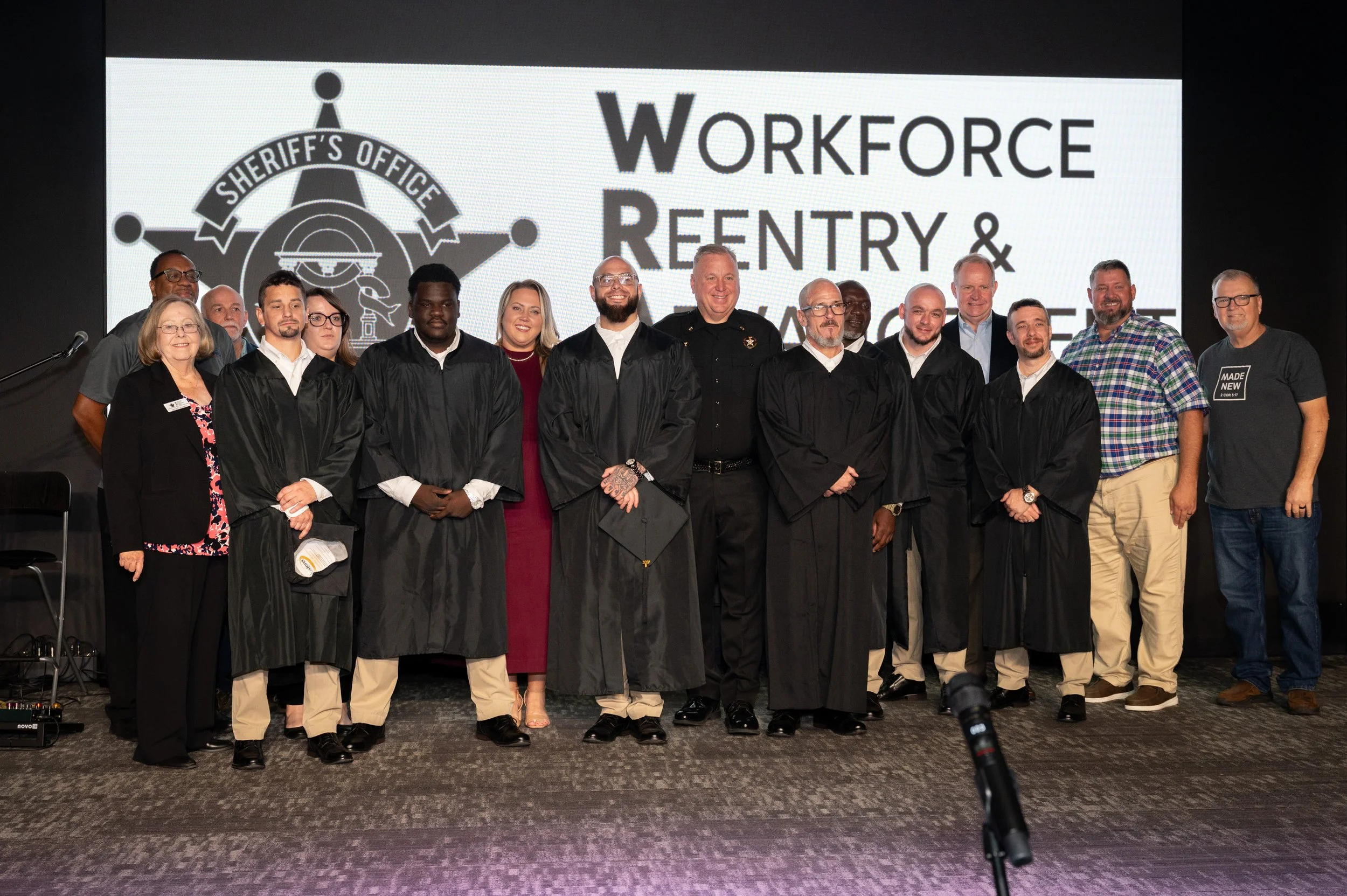 Group of people, including graduates in black robes, standing on stage in front of a large screen displaying a sheriff's badge and the words 'Workforce Reentry & Rehabilitation' at a formal event.