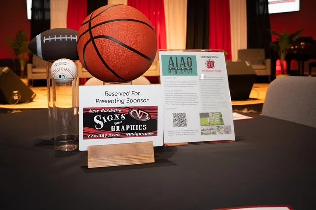 Basketball, football, and baseball on a table with sponsor signs and informational flyers at an indoor event.
