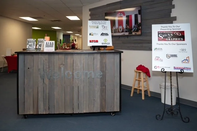 Reception desk with a TV screen mounted on the wall behind it, displaying a presentation with flags. To the right, a sign on an easel thank sponsors, with a stool and a red cloth on top. The reception desk has a rustic wood finish with the word 'Welc