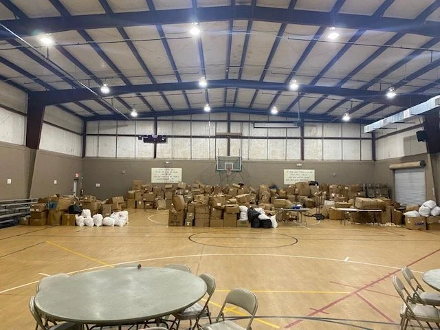 Indoor gymnasium filled with boxes and supplies, tables and chairs in the foreground.