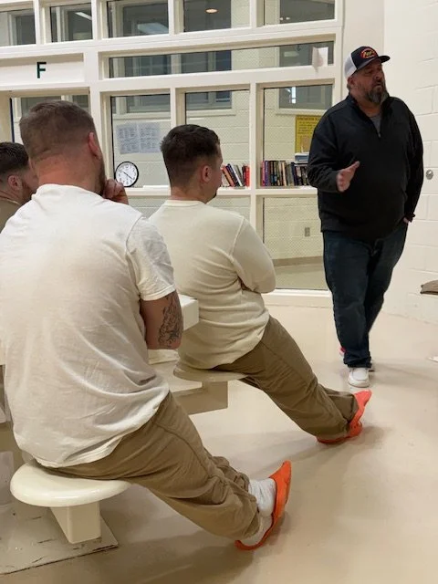 A man in a black hoodie and cap giving a presentation to three young men sitting on a bench in a room with glass windows and bookshelves.