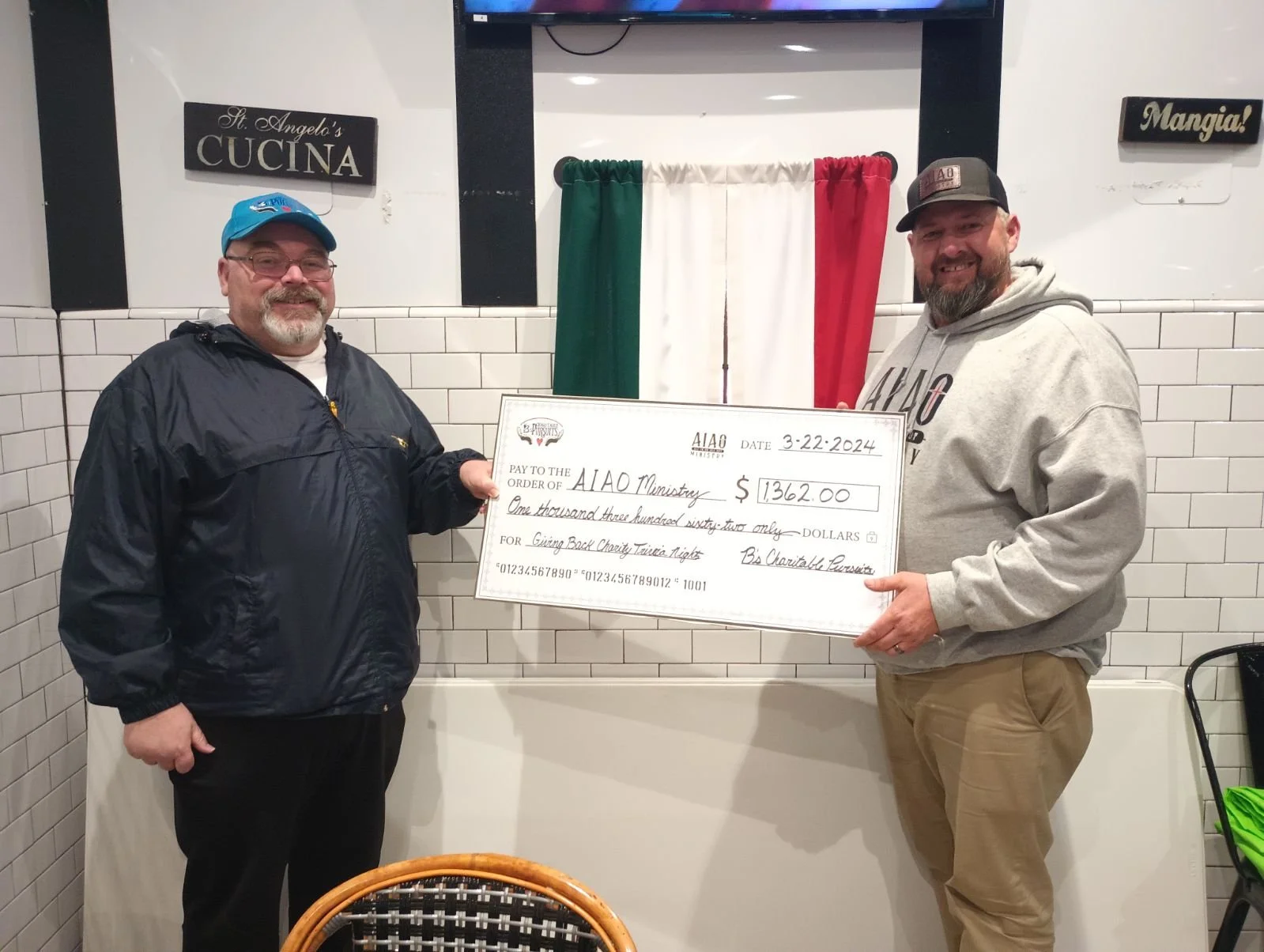 Two men standing in a restaurant holding an oversized check made out to AIao Ministry for $1,362.00. The man on the left wears glasses, a blue cap, and a black jacket, while the man on the right wears a gray hoodie and a cap. The background features 