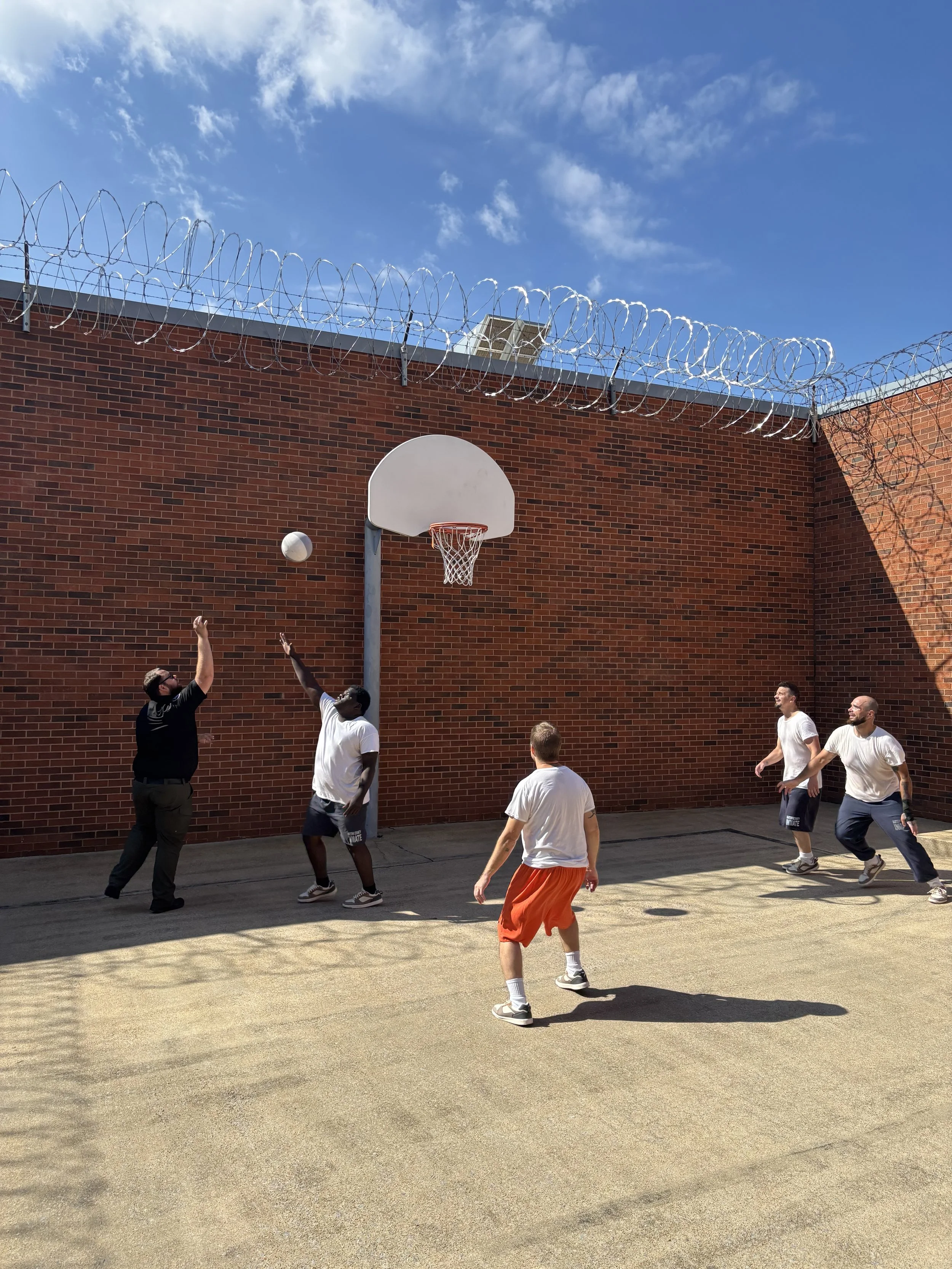 Group of five men playing basketball outdoors on a court with a brick wall, barbed wire, and blue sky in the background.