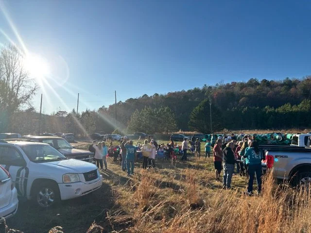 A large outdoor gathering of people in a grassy field with parked cars and trees in the background, under a clear blue sky with the sun shining brightly.