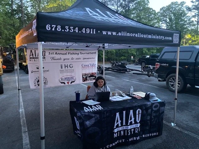 A woman sitting at a portable table under a black canopy tent with 'All In or All Out Ministry' branding. The table has a laptop, water bottle, papers, and a sun hat. In the background, a banner displays sponsorship information for the 1st Annual Fis