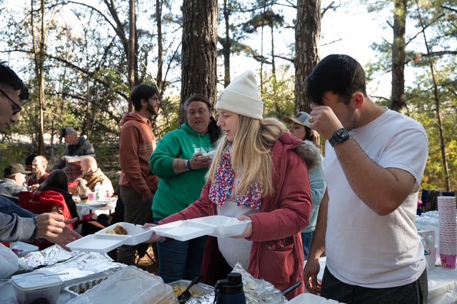 People gathered outdoors in a wooded area, serving food from a table with trays and containers, during what appears to be a community event or picnic.