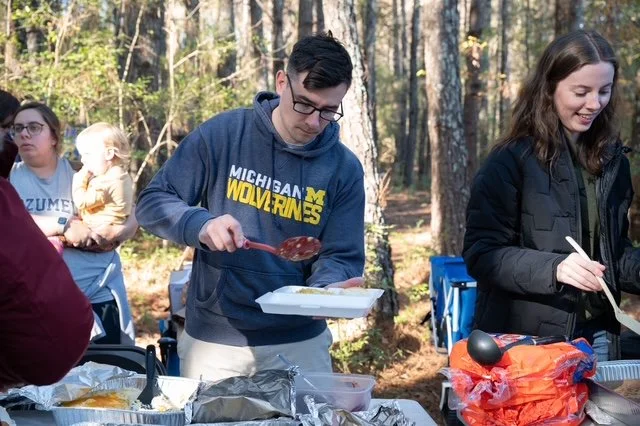 People outdoors at a park with food, including a young man in a Michigan Wolverines hoodie serving himself from a tray and a young woman in a black jacket eating, with other individuals and trees in the background.