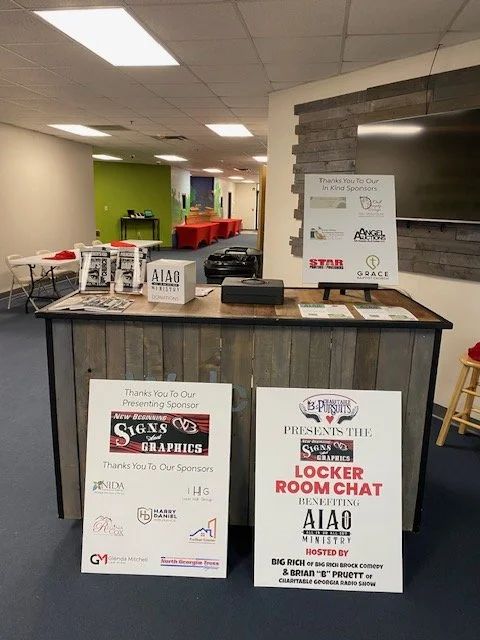 Event space with a registration or information desk in the foreground, featuring signs promoting sponsor acknowledgments and a charity event, with tables and chairs in the background.