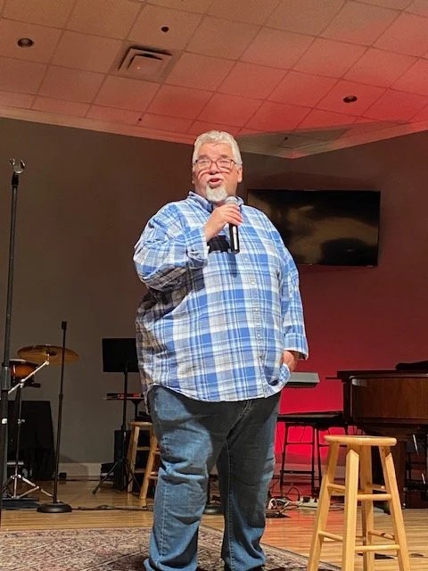 A man with gray hair and glasses, wearing a blue plaid shirt, speaking into a microphone on stage in a room with musical instruments and a television mounted on the wall.