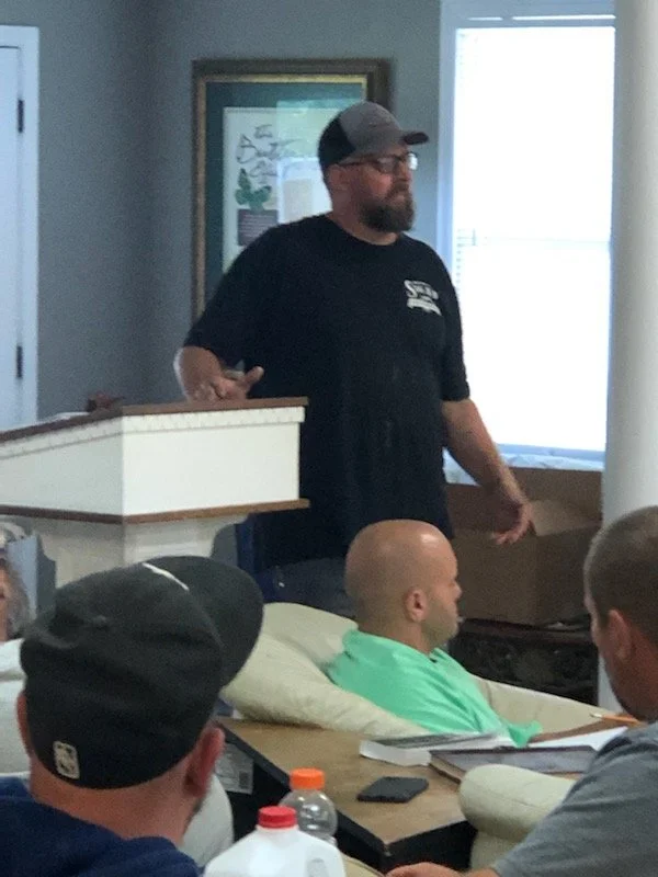 A man with beard, glasses, and a cap standing behind a table, speaking to a group of people seated at a meeting or classroom setting.