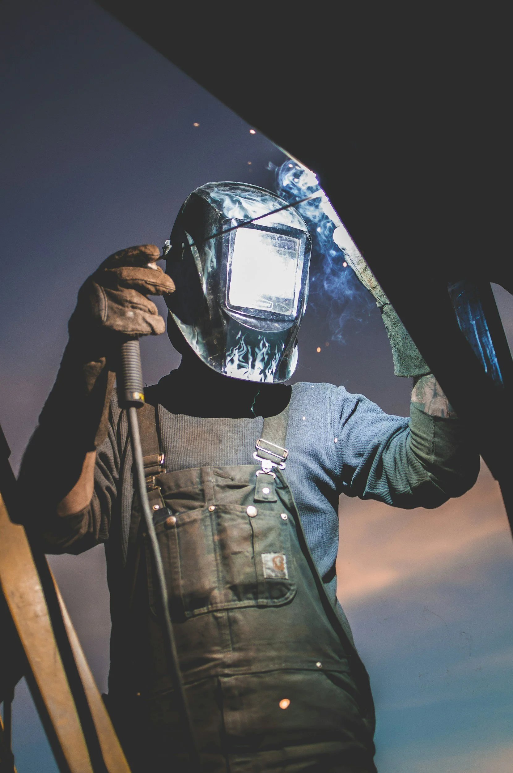 A welder wearing protective gloves, a welding helmet with a visor, and overalls, working outdoors during dusk or night, with sparks and bright light from welding.