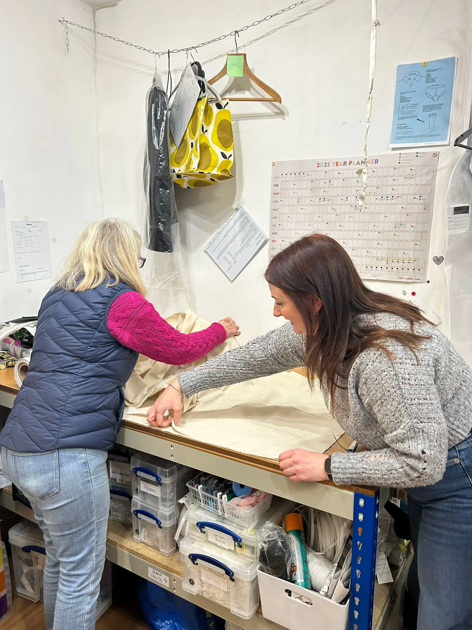Two women working together in a cluttered workshop or storage room, sorting or preparing materials on a worktable. Behind them are shelves with labeled bins and various supplies such as tape, scissors, and paper.