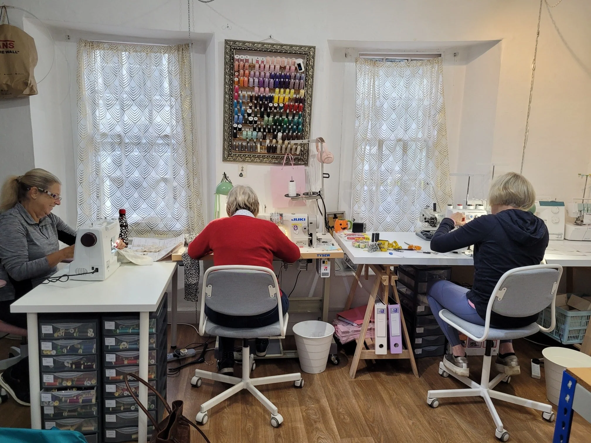 Three women sitting at tables working on sewing projects in a well-lit room with white walls and window curtains. The room has sewing machines, storage drawers, and sewing supplies.