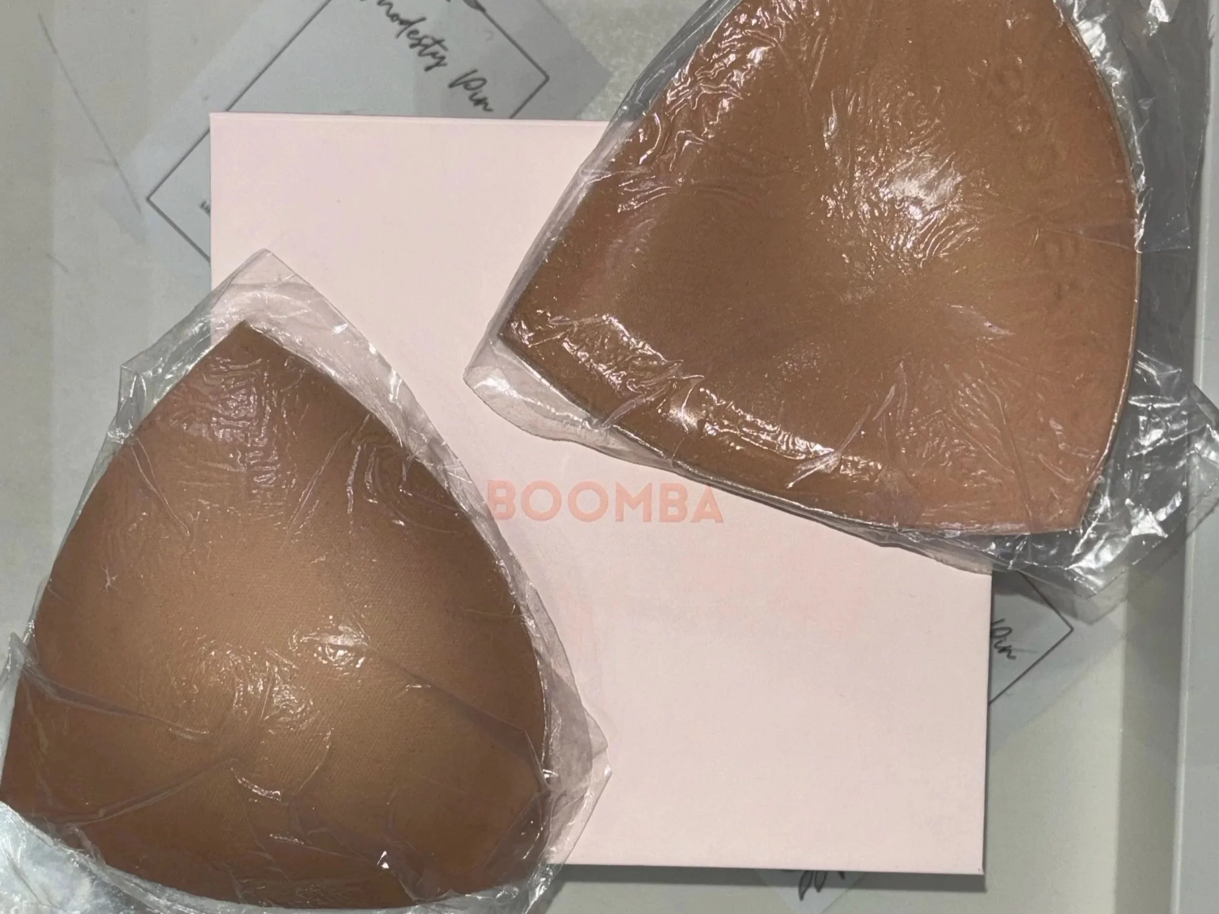 Two square pieces of brown fudge, individually wrapped in clear plastic, placed on a white box labeled "BOOMBA". Some papers and a gray tile surface are also visible in the background.