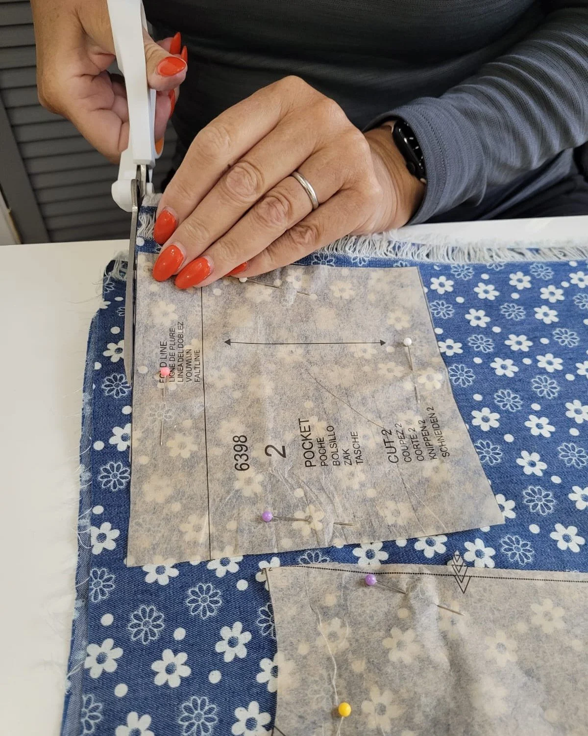 A person sewing fabric using scissors and pins on a blue and white floral fabric, with a pattern pattern paper labeled in multiple languages.
