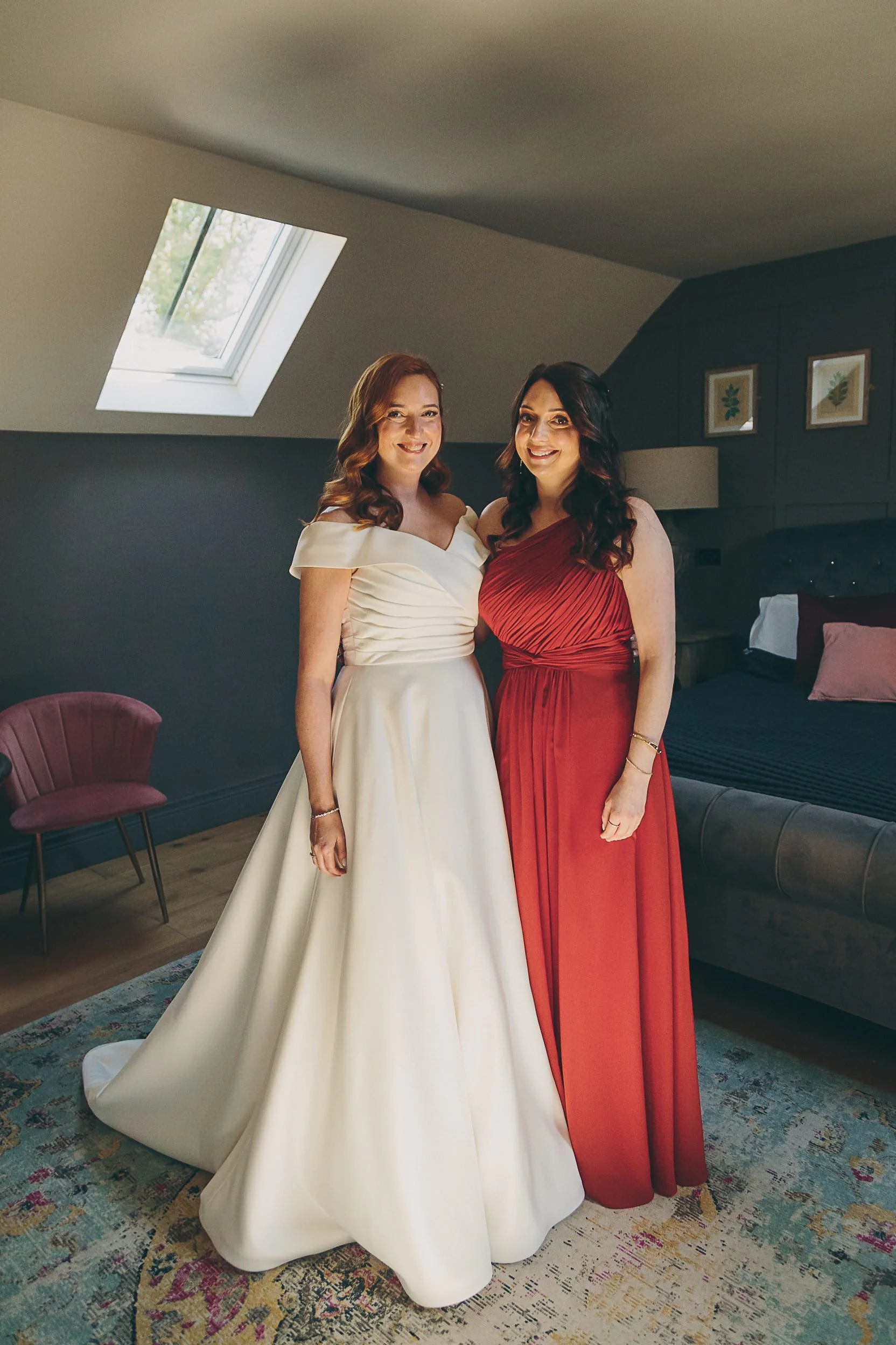 Two women in formal dresses smiling indoors, one in a white gown and the other in a red gown, standing on a patterned carpet in a room with dark walls and framed pictures, with a skylight on the ceiling.