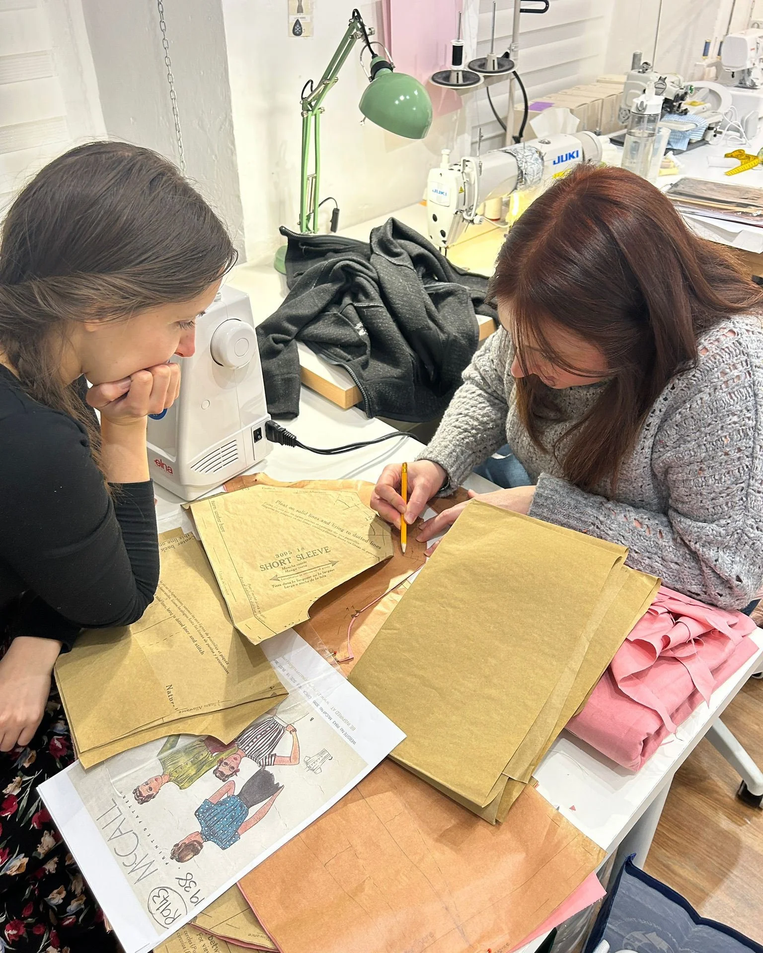 Two women working on fashion design projects at a cluttered workspace, with fabric swatches, pattern papers, and a sewing machine.