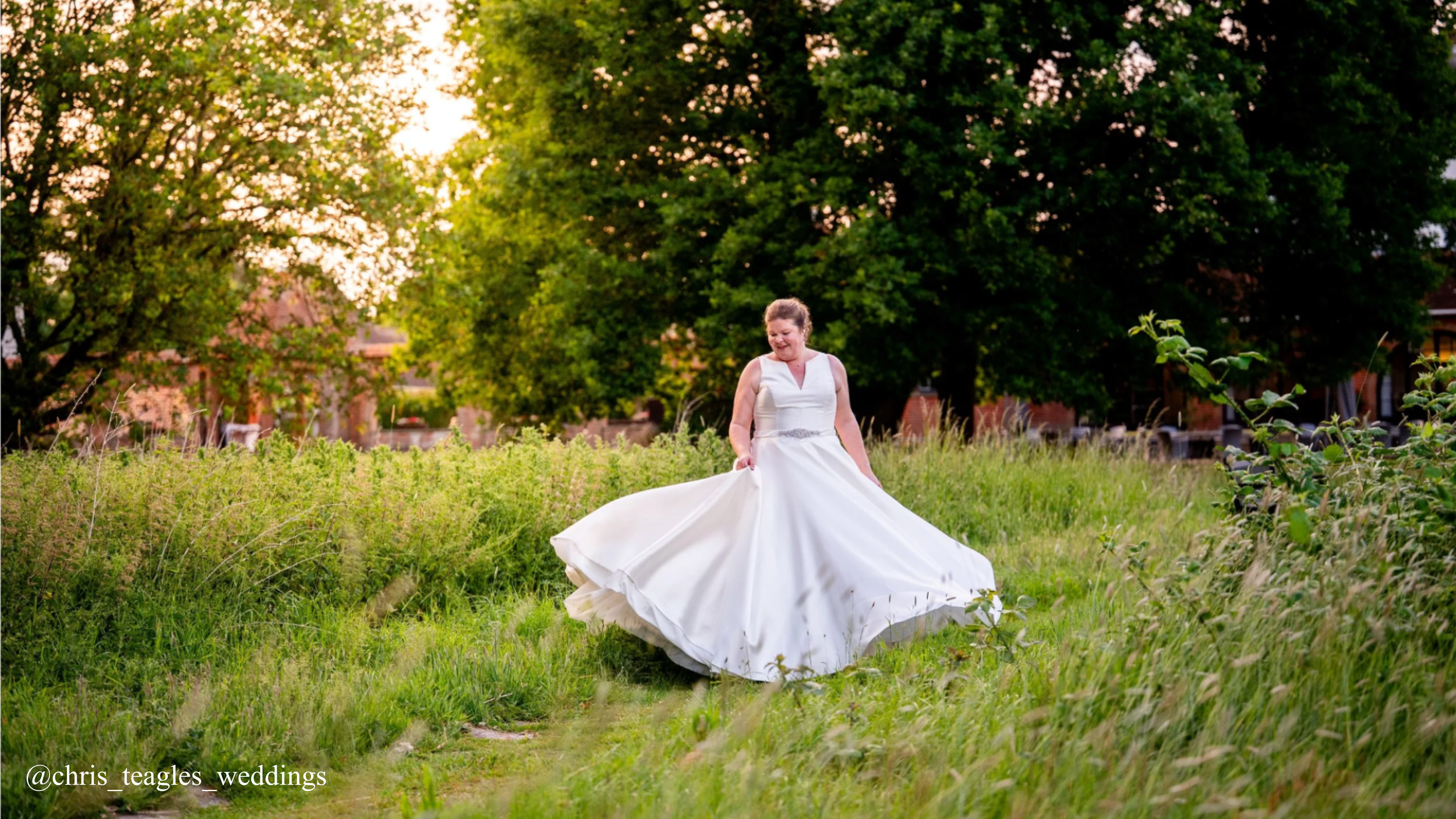 A bride in a white wedding dress is standing in a grassy field, smiling and lifting her dress slightly, with large green trees and sunlight in the background.