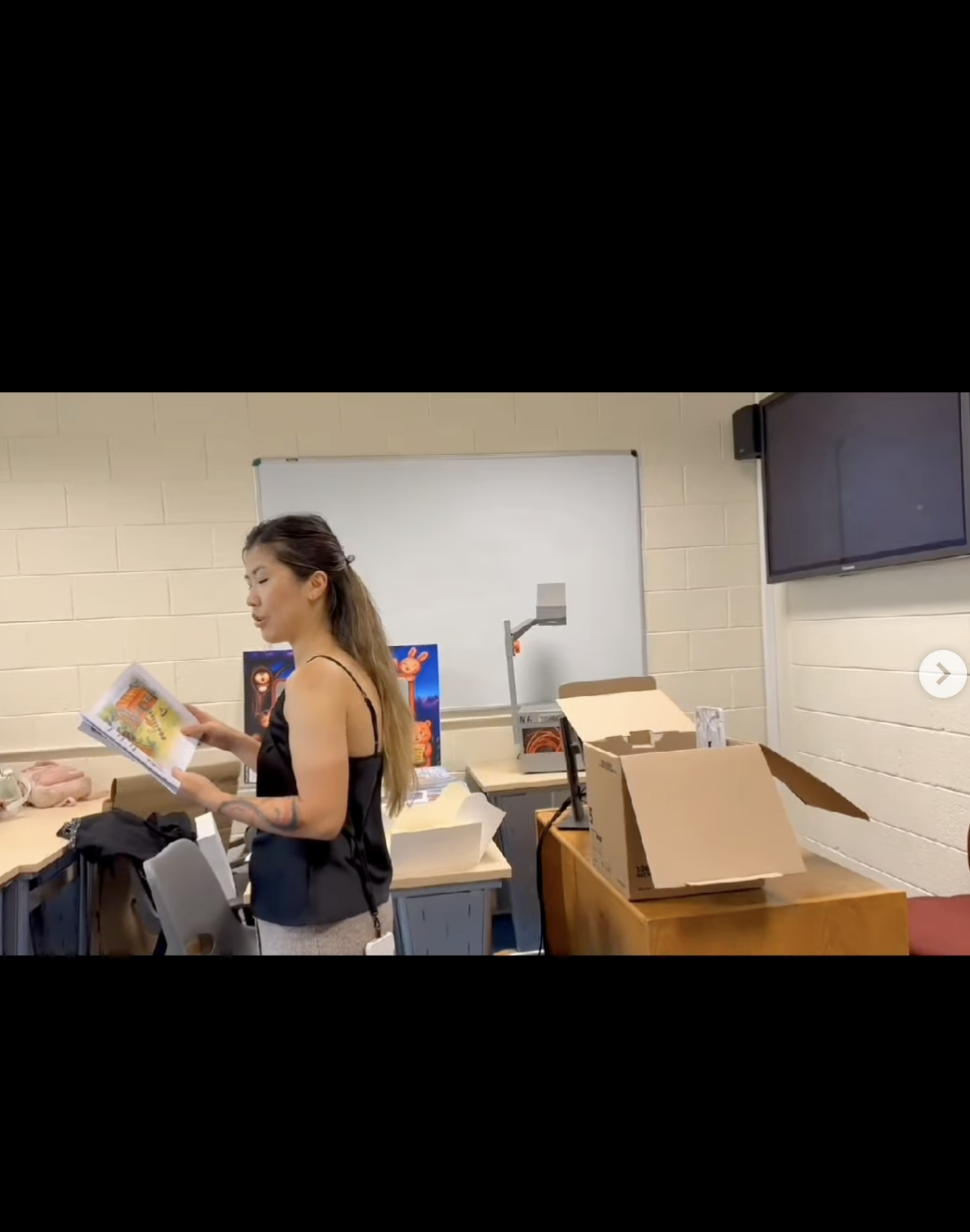 A woman in a black sleeveless top and gray pants standing in a classroom or office, holding and looking at an open magazine, with various items like cardboard boxes and a whiteboard in the background.