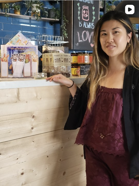 A young woman with long hair standing next to a wooden counter in a cafe, with shelves of packaged products and a blackboard behind her.