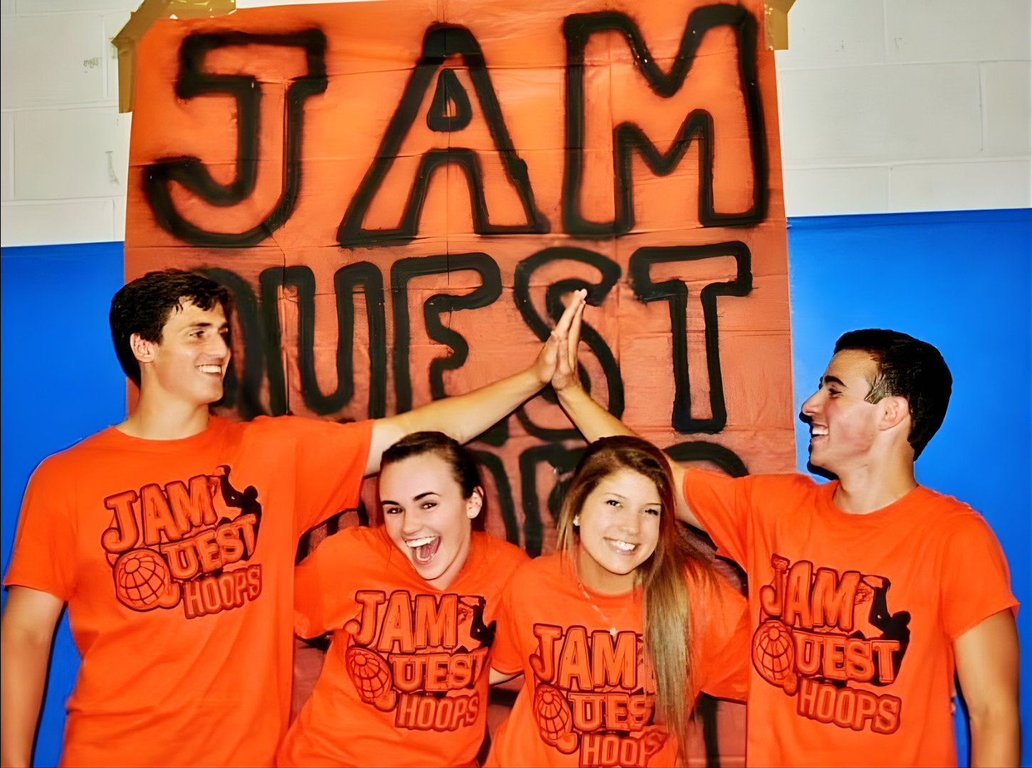 Four teenagers wearing matching orange T-shirts with 'JAM QUEST HOOPS' logo, giving each other high fives in front of a large banner with the words 'JAM' and 'QUEST' in black spray paint on an orange background. The scene appears to be at a basketball event or competition.