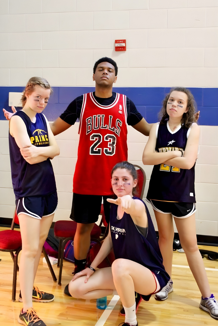 Four young girls in basketball uniforms posing with a teenage boy in a gymnasium. The boy is wearing a red Bulls jersey with the number 23, and the girls are in navy and black jerseys with the word 'Prince' on them. One girl is sitting on the floor pointing at the camera, while the others stand with confident expressions.