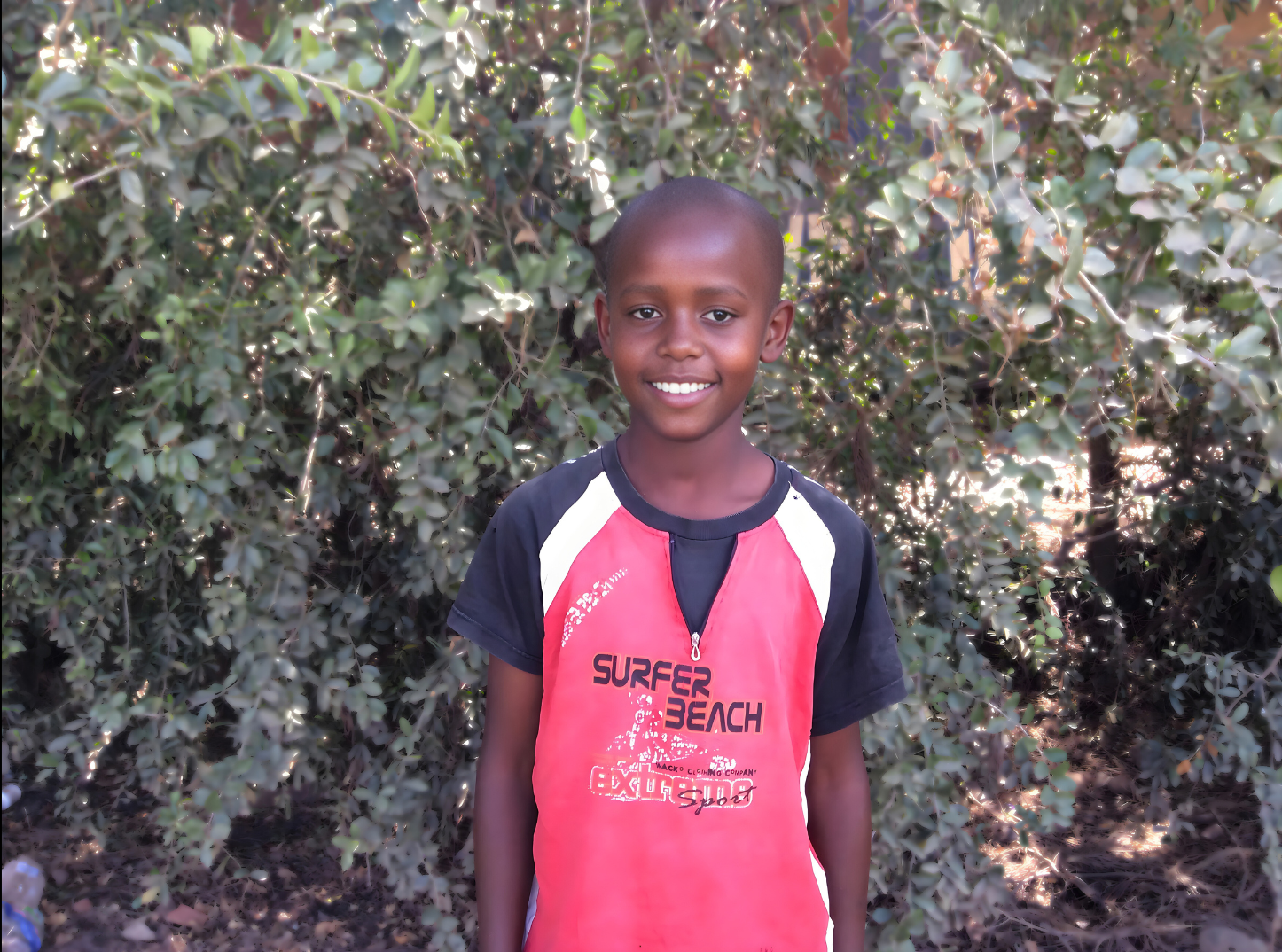 A young boy standing outdoors in front of green bushes. He is smiling, has short hair, and is wearing a red t-shirt with black and white accents that says 'Surfer Beach'.