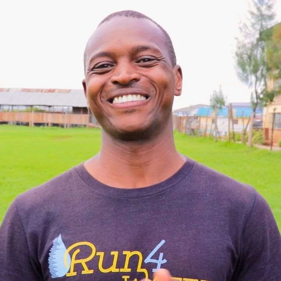 Smiling man outdoors in a park with green grass, trees, and buildings in the background, wearing a black T-shirt with yellow text.