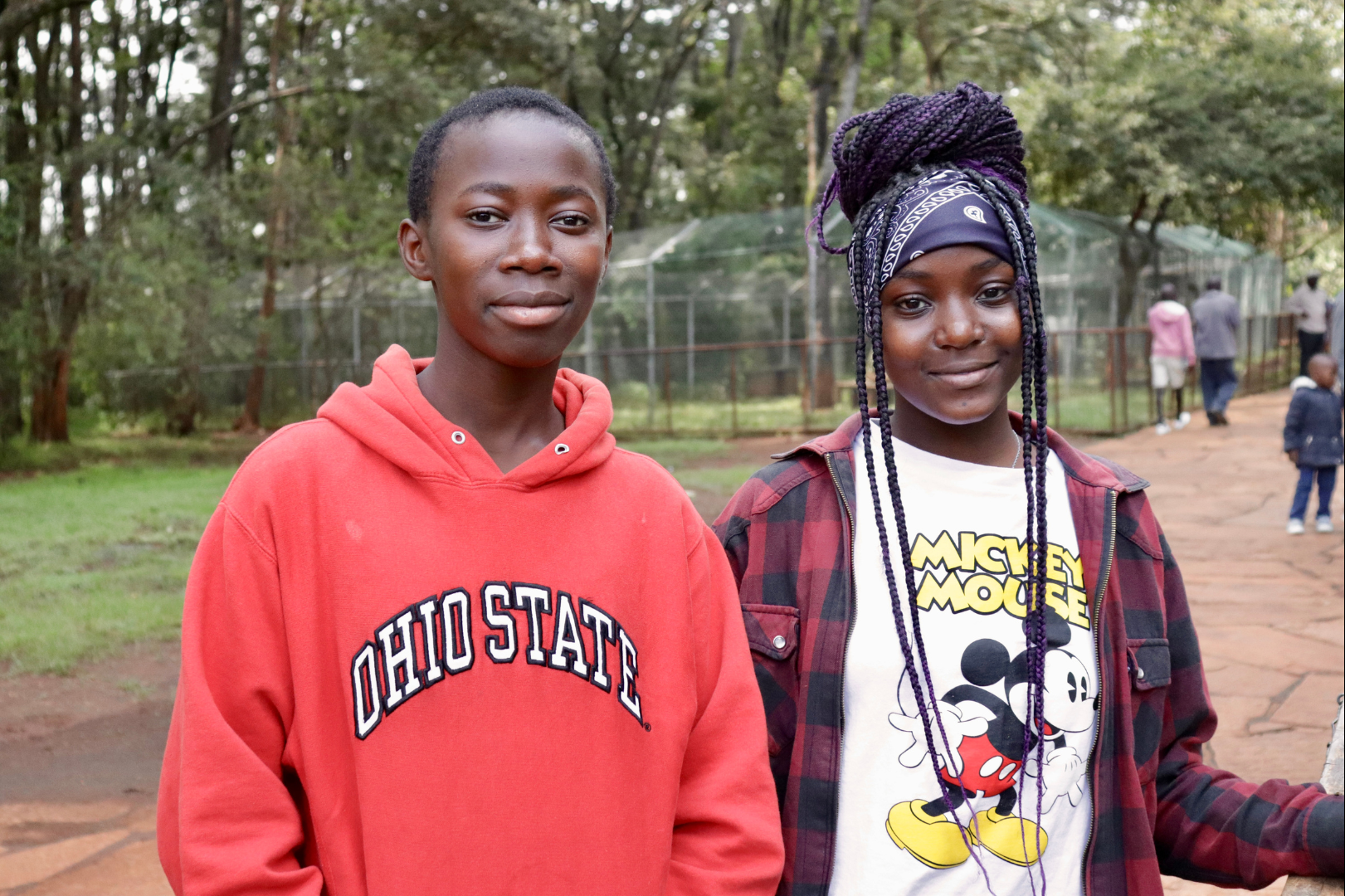 Two young women standing outdoors in a park or zoo, with trees and a fence in the background. One is wearing a red Ohio State hoodie, and the other is wearing a Mickey Mouse t-shirt with a plaid jacket and a bandana in her hair.