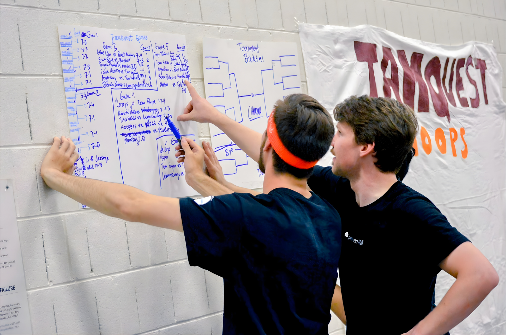 Two young men with black t-shirts and one with a red headband discussing a tournament bracket and game schedule written on white poster papers on a wall, with a large banner that reads 'TROPHY BOO' in the background.