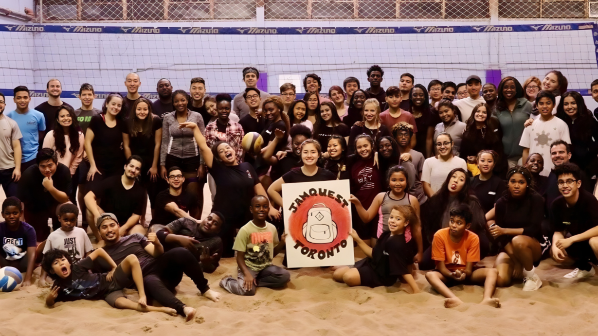 A large group of people, including children, teenagers, and adults, gathered on an indoor volleyball court for a group photo. Some are holding volleyballs, and there is a person in the center holding a sign that says 'Tamqueet Toronto' with a backpack illustration. The background features a volleyball net and a sports hall setting.