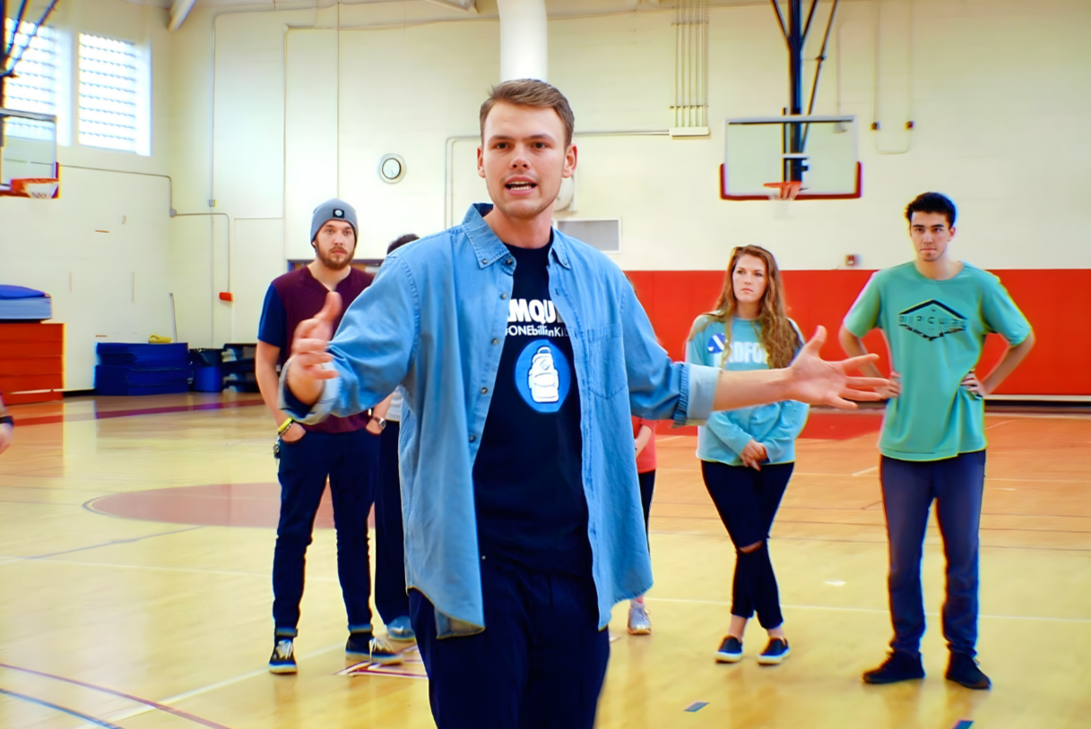 A young man in a blue shirt speaking or instructing a group of teenagers in a gymnasium.