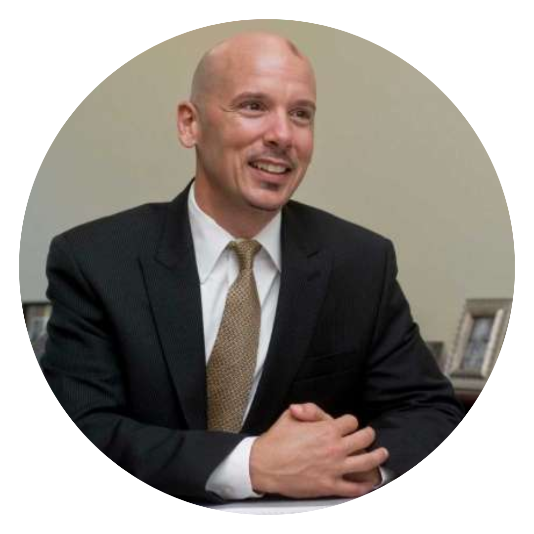 A bald man in a suit and tie smiling, sitting at a desk with framed photos in the background.