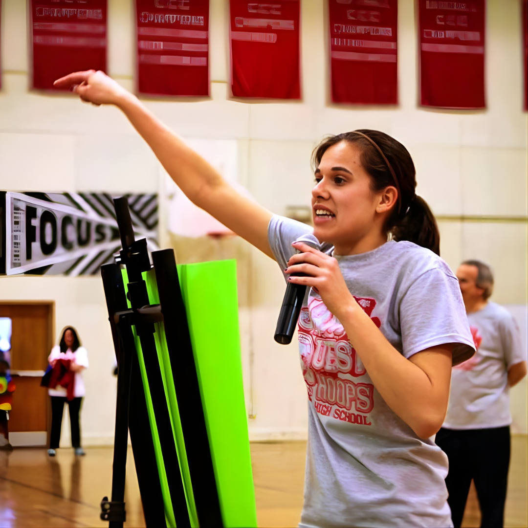 A young woman with brown hair, wearing a gray T-shirt, is speaking into a microphone and gesturing with her right arm during a presentation or speech in an indoor gymnasium or hall. There are banners and other people in the background.