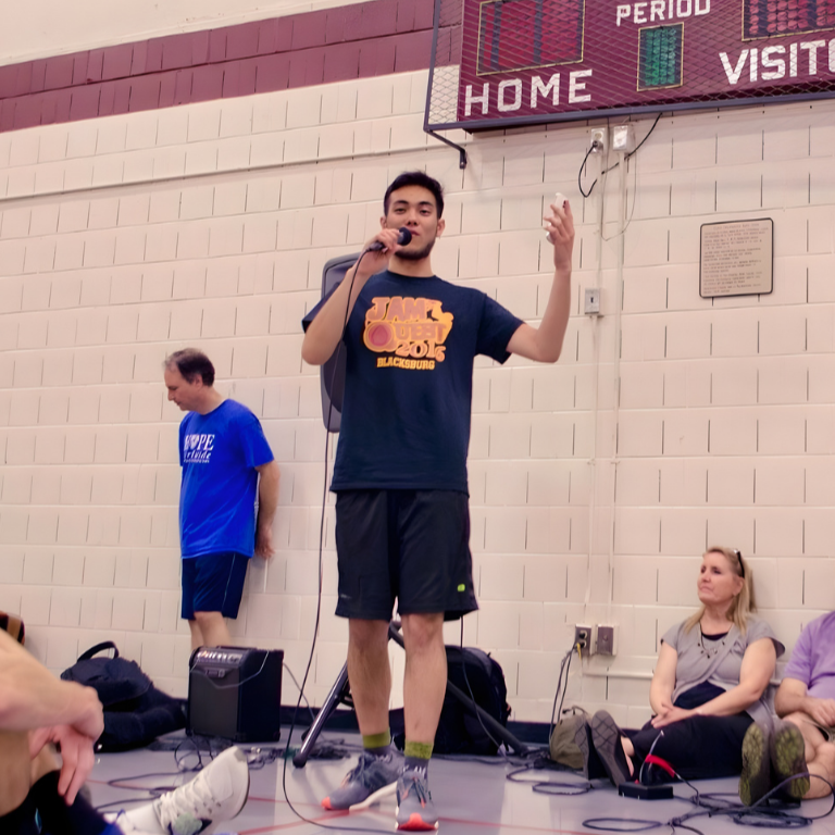 A young man speaking into a microphone on a gymnasium floor with a basketball scoreboard in the background, while two people sit on the floor and others stand nearby.