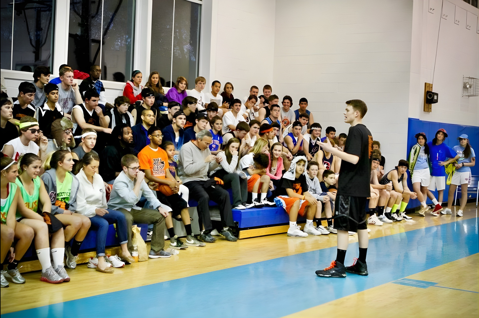 A young man in a black T-shirt and shorts giving a speech to a large audience of young basketball players and spectators in a gymnasium.
