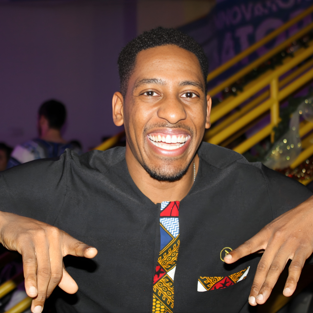 A man smiling broadly at a social event, wearing a black shirt with colorful patterned design on the front pocket and shirt placket, gesturing with his hands, background with yellow stairs and purple lighting.