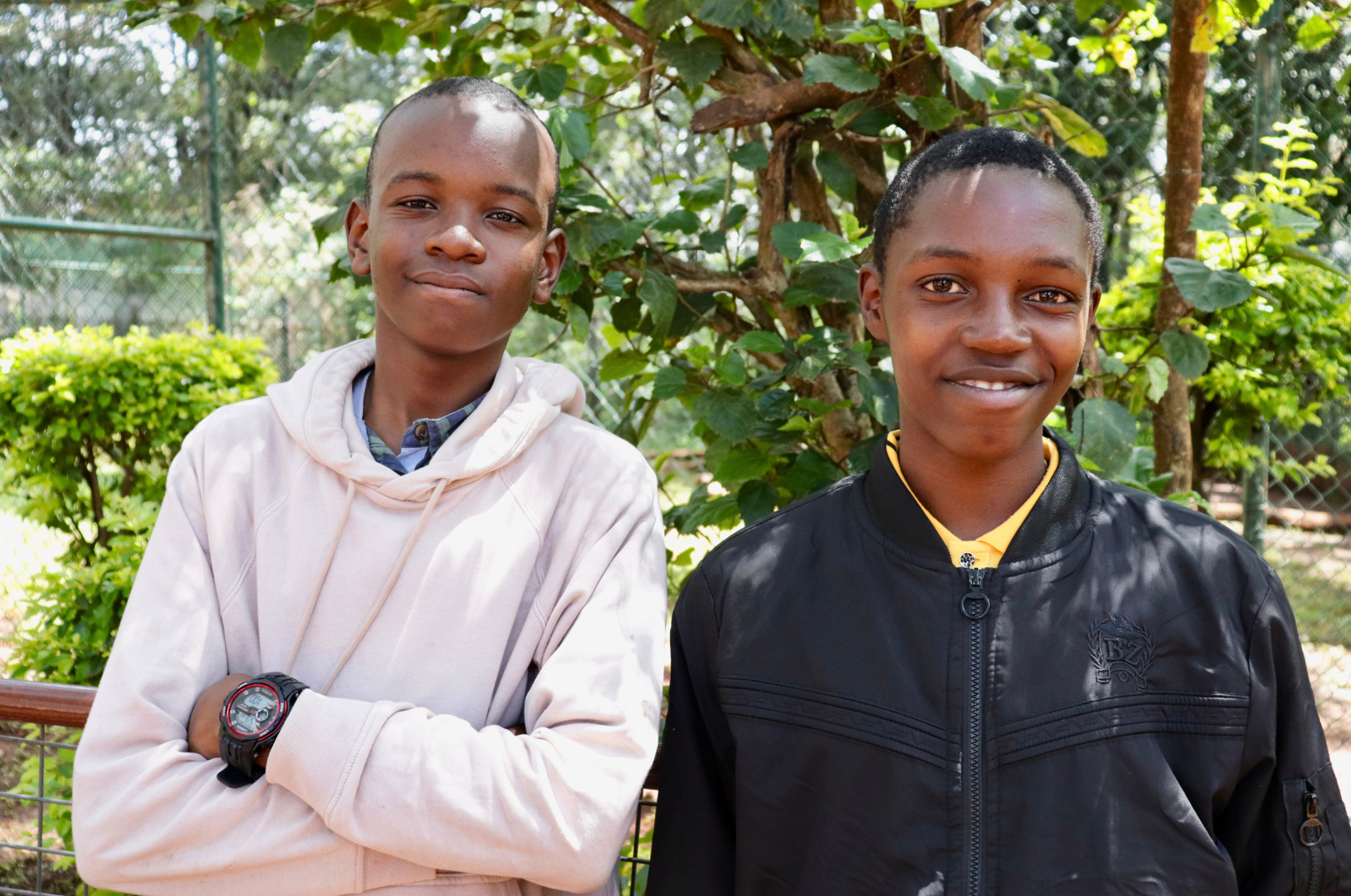 Two boys standing outdoors in front of green foliage and a fence, smiling at the camera.