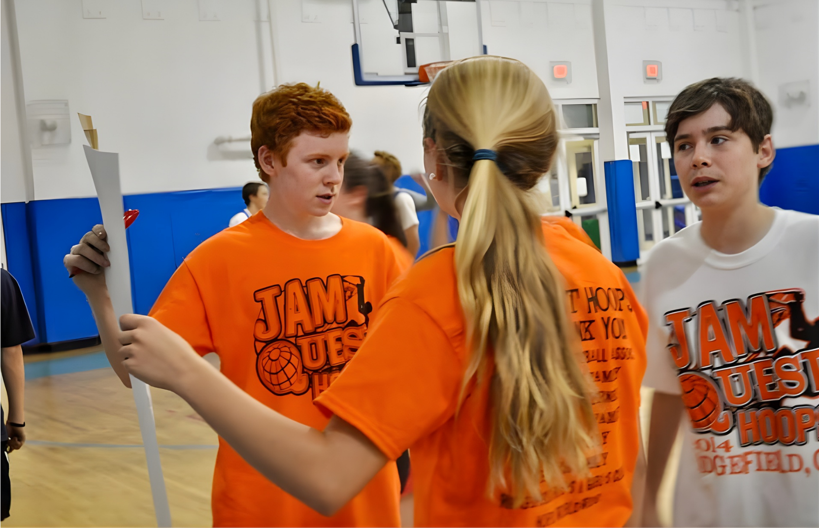 Three teenagers inside a gymnasium having a conversation. All are wearing T-shirts with 'JAM TUESDAY HOOP' and a basketball logo. The background shows a basketball hoop and other young people.