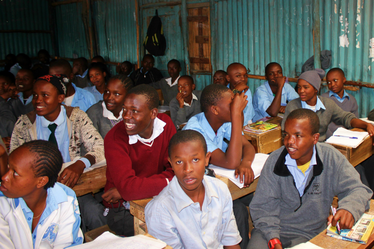 Young students in a classroom with tin walls, sitting at wooden desks, engaging and smiling.
