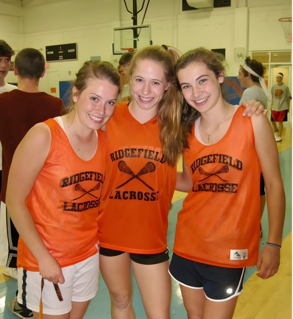 Three young women in orange lacrosse jerseys with 'RIDGEFIELD LACROSSE' and crossed lacrosse sticks printed on them, smiling and standing together in a gymnasium.
